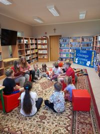 Dzieci siedzą w kółku w bibliotece szkolnej. Dziewczynki  z klasy szóstej czytają innym książkę
