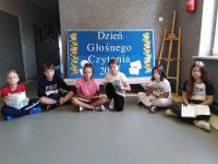 Uczniowie klasy piątej siedzą w korytarzu szkolnym na podłodze i czytają książki. Za nimi dekoracja