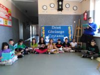 Uczniowie klasy czwartej siedzą w korytarzu szkolnym na podłodze i czytają książki. Za nimi dekoracja