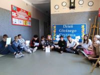 Uczniowie klasy ósmej siedzą w korytarzu szkolnym na podłodze i czytają książki. Za nimi dekoracja