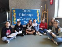 Uczniowie klasy szóstej siedzą w korytarzu szkolnym na podłodze i czytają książki. Za nimi dekoracja