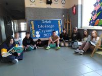 Uczniowie klasy siódmej siedzą w korytarzu szkolnym na podłodze i czytają książki. Za nimi dekoracja