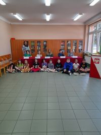 Na podłodze siedzą uczniowie klasy czwartej, za nimi na krzesłach przy stole siedzi komisja uczniowska. Obok stoi nauczyciel