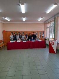 Na krzesłach za stołem siedzi komisja uczniowska, za nimi stoją uczniowie z klasy siódmej