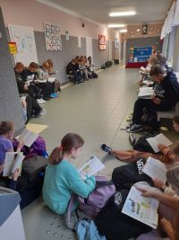 Uczniowie i nauczyciele siedzą w korytarzu szkolnym i czytają książki