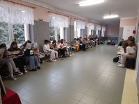 Uczniowie i nauczyciele siedzą w korytarzu szkolnym i czytają książki