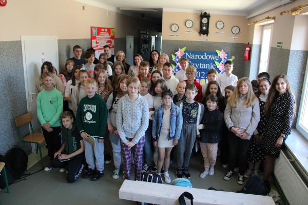 Uczniowie wraz z nauczycielami stoją w grupie, w tle dekoracja Narodowe Czytanie